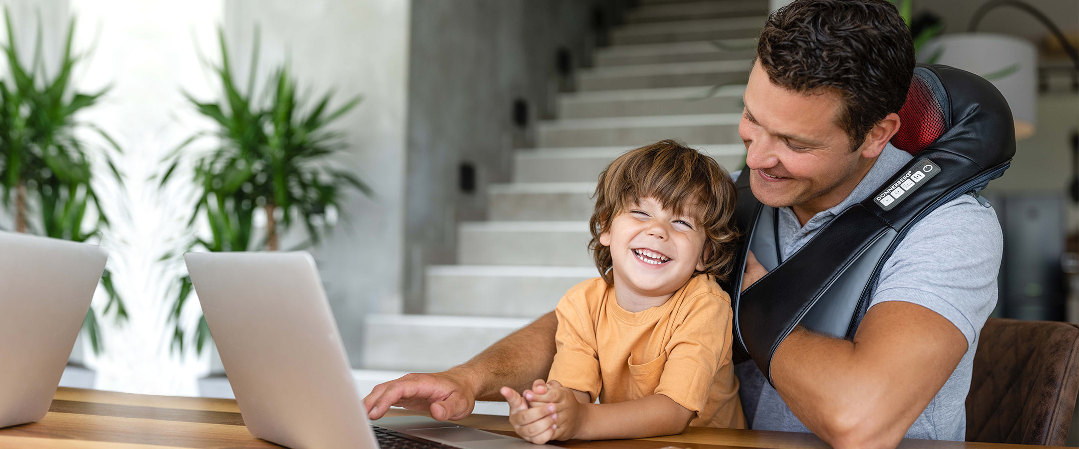 dad and son playng on laptop while dad relaxes with neck massager